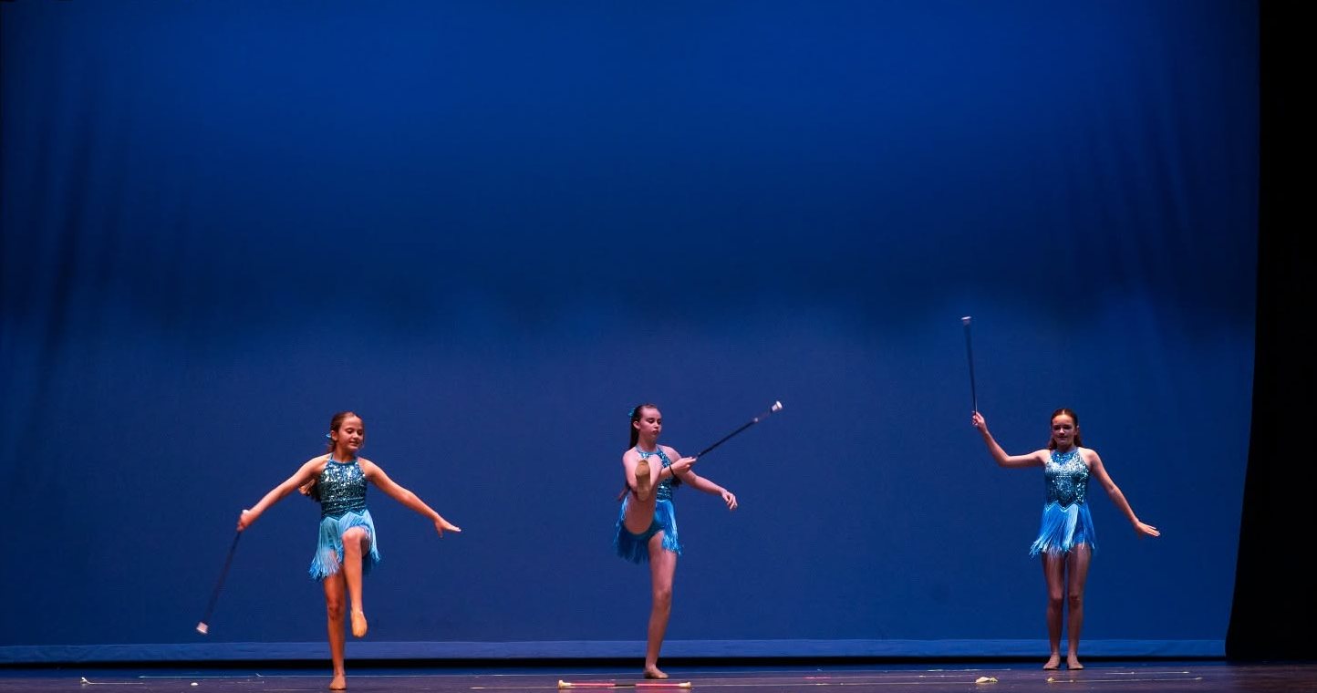 Three baton twirlers in turquoise fringe costumes perform on a blue-lit stage