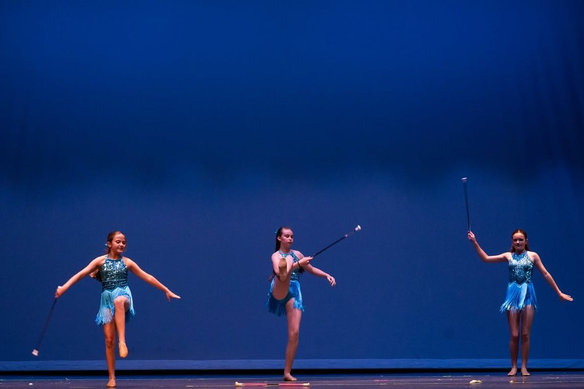 Three baton twirlers in turquoise fringe costumes perform on a blue-lit stage