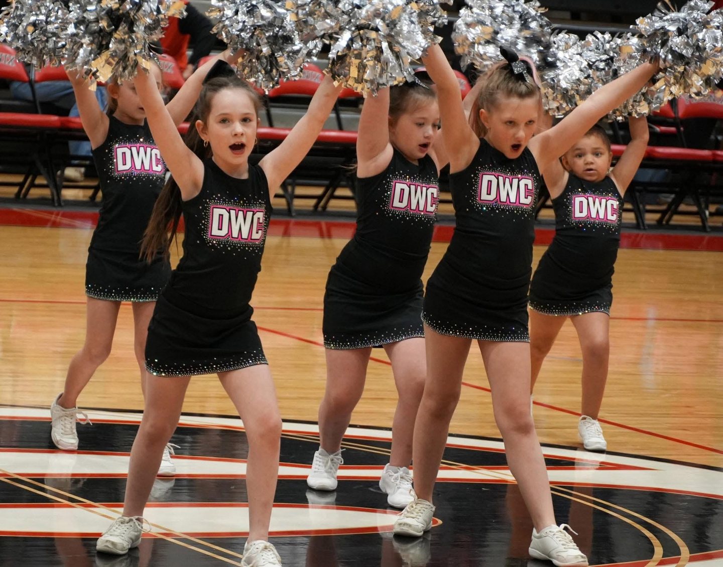 Young cheerleaders in black DWC uniforms raise silver poms overhead on a basketball court