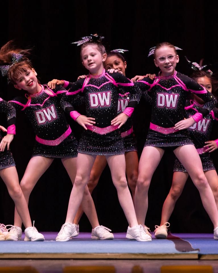 Cheerleaders in black and pink DW uniforms with sparkle bows pose with linked arms on a stage