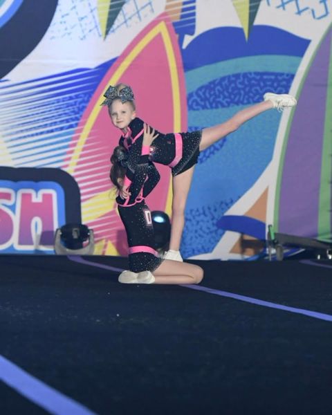 Two young cheerleaders in black and pink sparkle uniforms on a stage, one kneeling while the other holds a leg extension