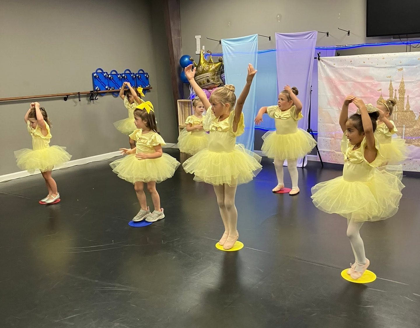 Young dancers in yellow tutus raise their arms on colored floor spots in a dance studio