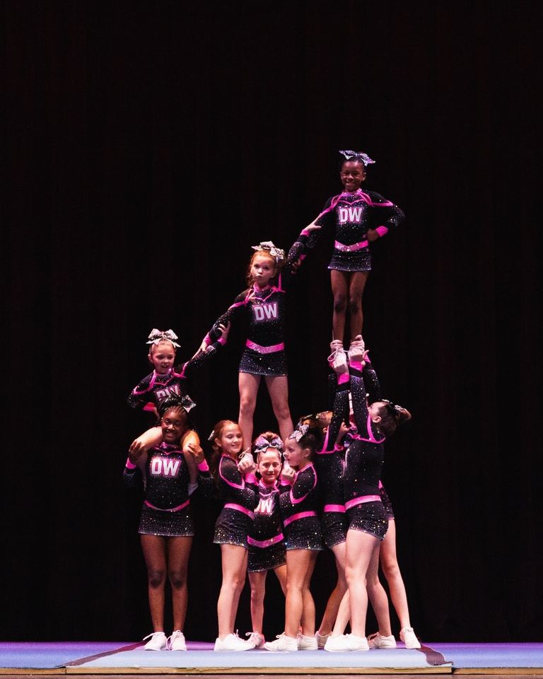 A youth cheer team in black and pink sparkle uniforms builds a pyramid with two flyers on a dark stage