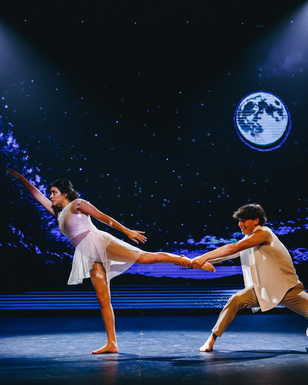 A dancer in a white dress extends into an arabesque while her partner holds her foot, under a projected moon and starry sky