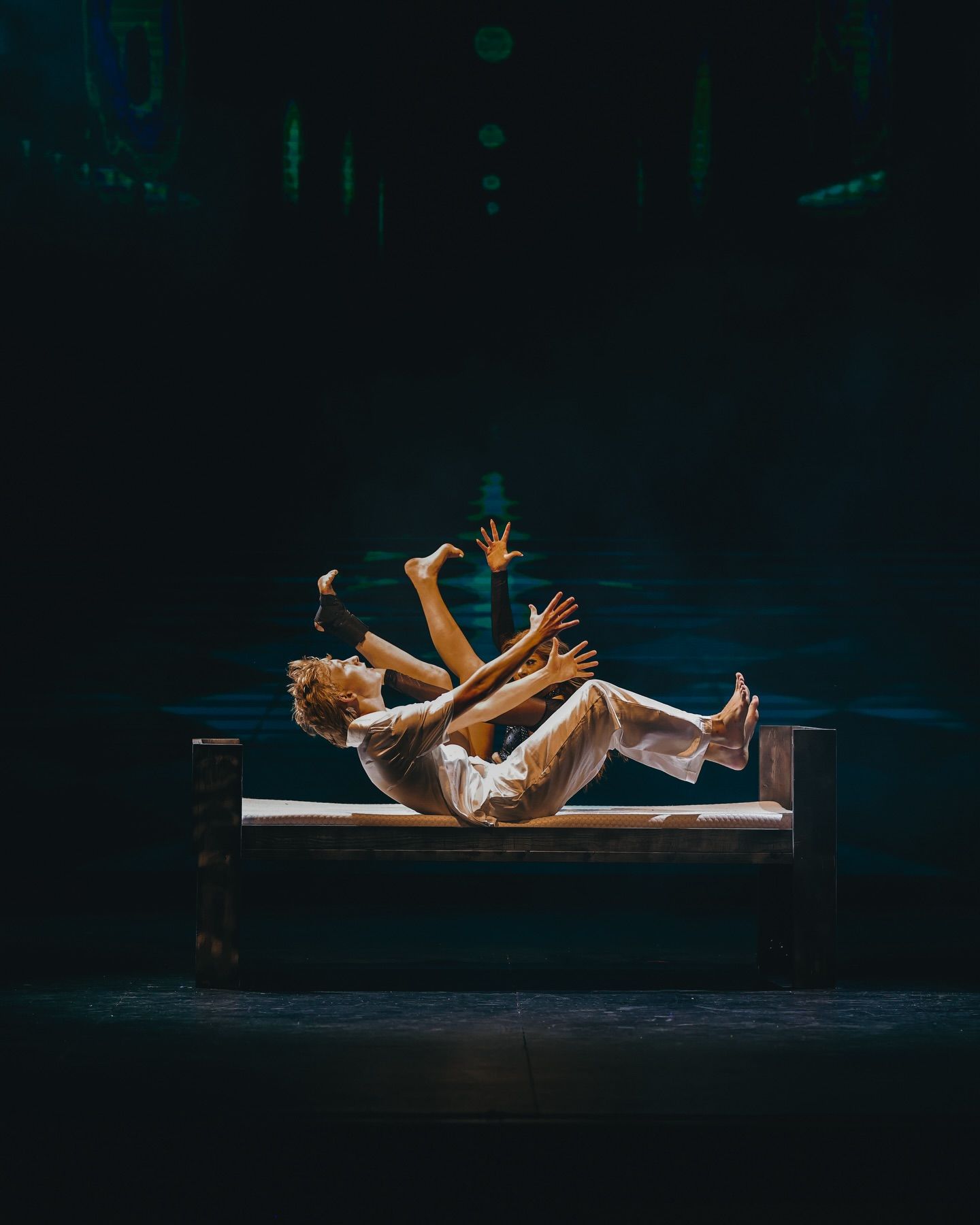 Two dancers on a wooden bench under a spotlight, one falling back with arms reaching up, against a dark teal stage