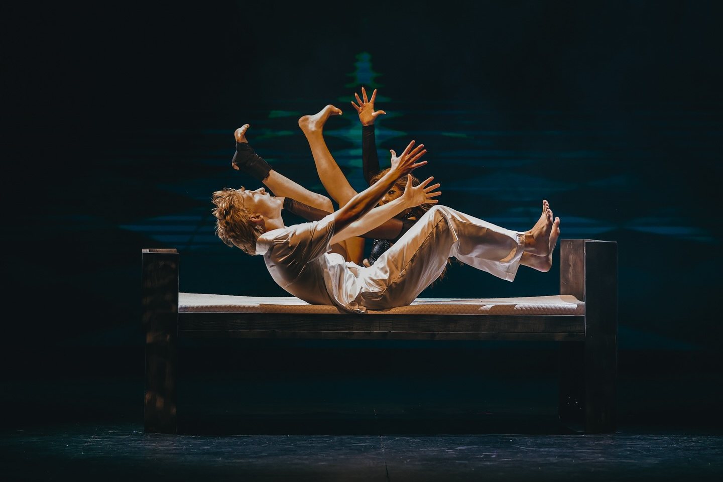 Two dancers on a wooden bench under a spotlight, one falling back with arms reaching up, against a dark teal stage