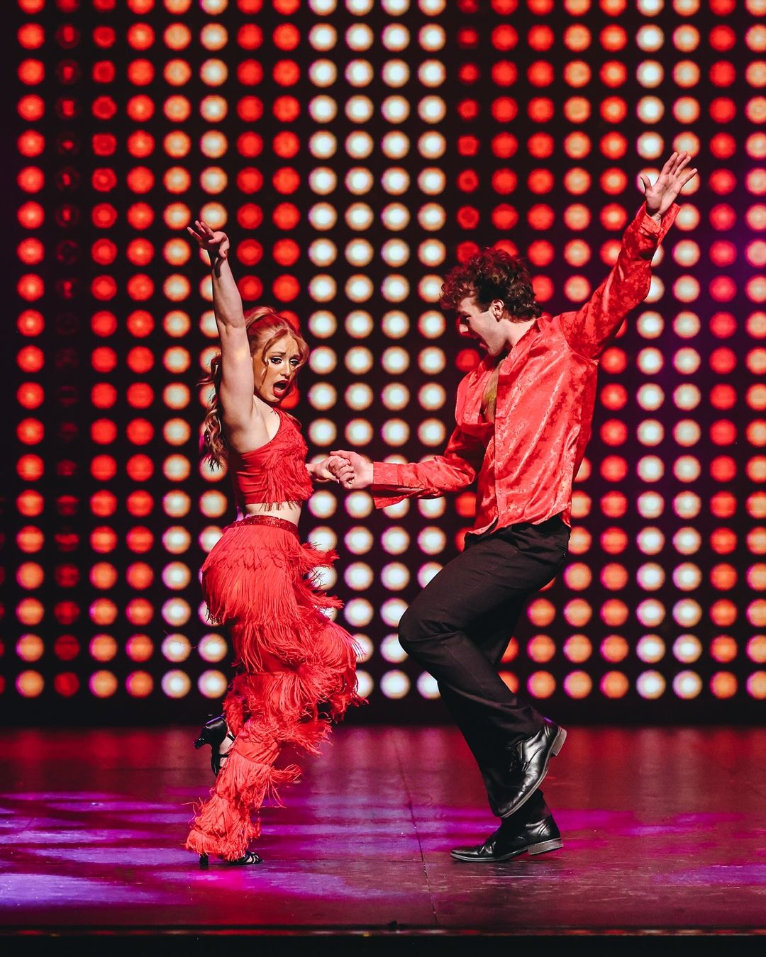 Two dancers in red fringe and satin costumes dance with arms raised in front of a glowing wall of stage lights