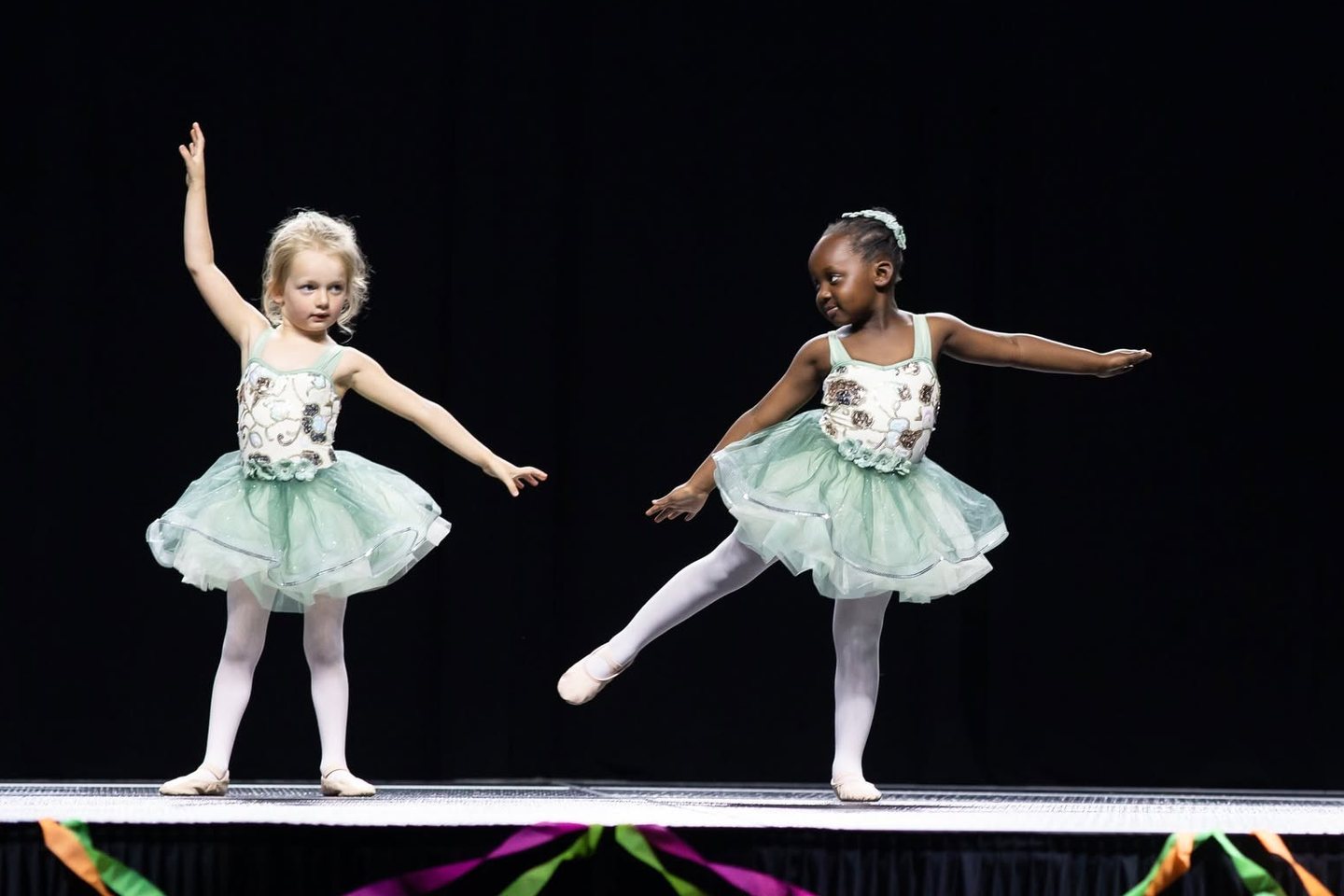 Two young dancers in mint tulle tutus pose with arms out on a stage edged with ribbons