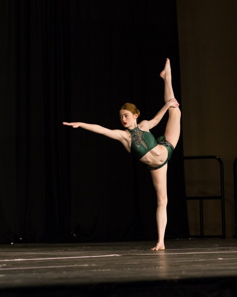 A teen dancer in a green costume holds one leg high in a standing extension on a dark stage