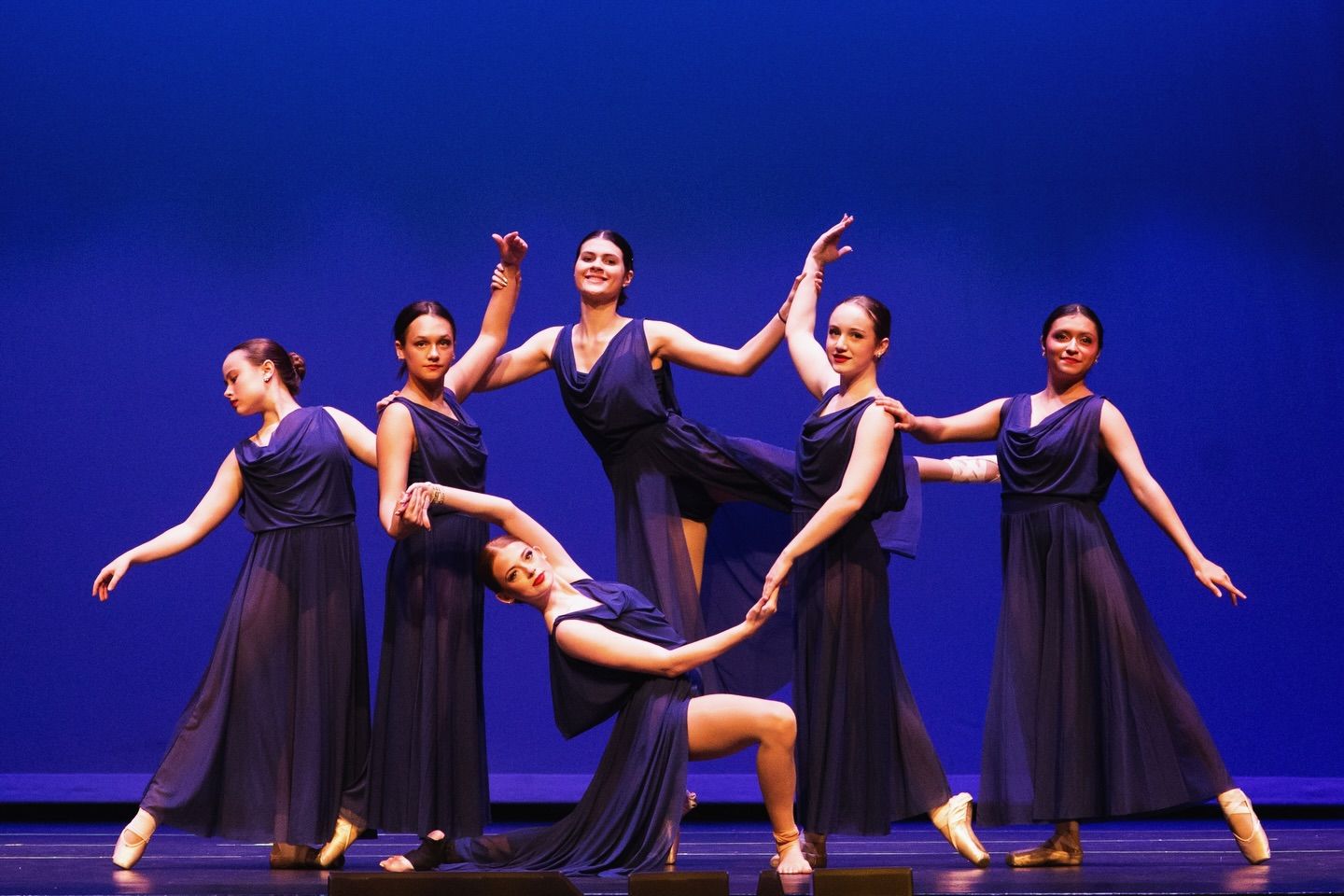 Six dancers in navy chiffon dresses and pointe shoes pose together on a stage lit deep blue