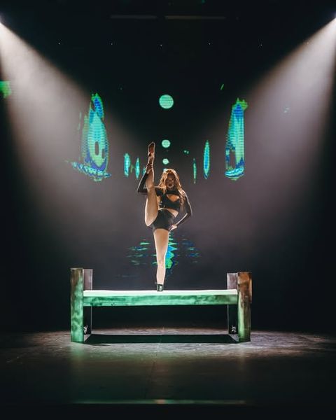 A dancer holds a standing high kick on a bench under two spotlights on a dark stage