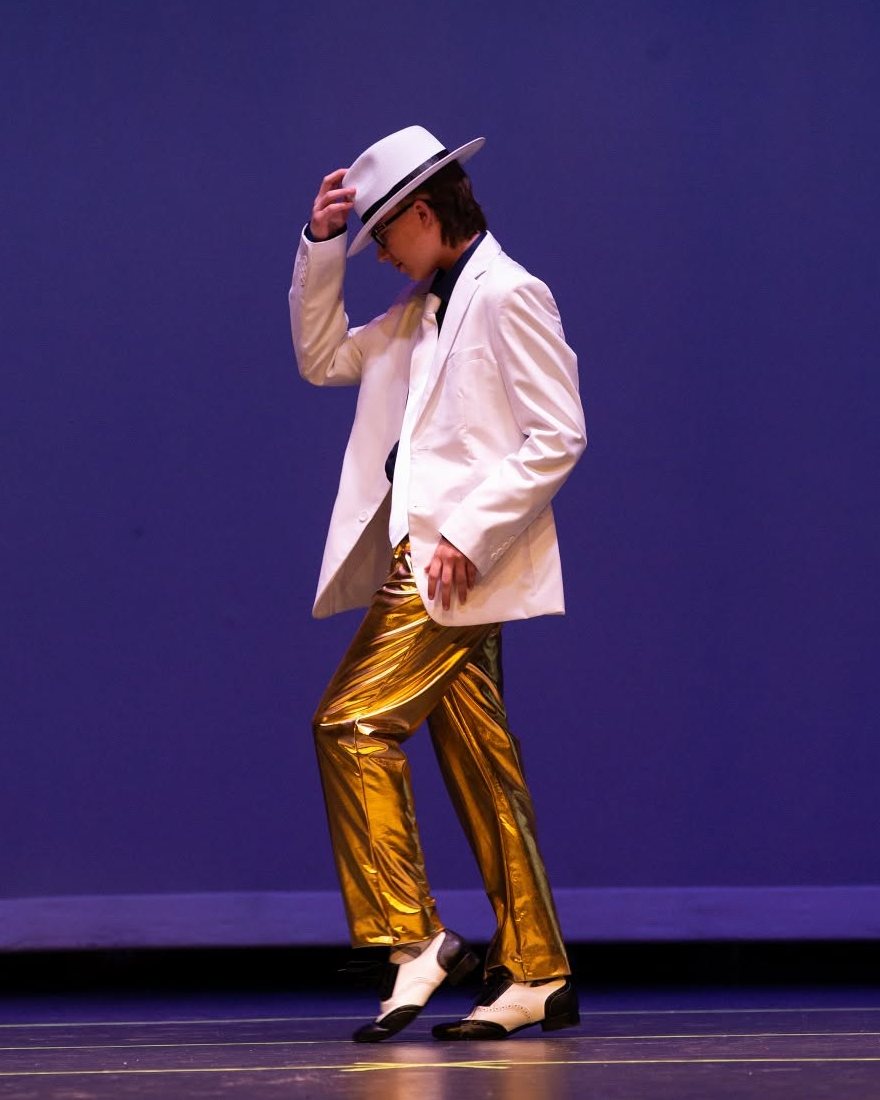 A dancer in a white suit, white hat and gold trousers tips the hat on a purple-lit stage