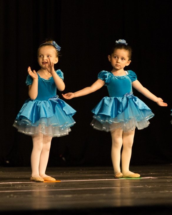 Two dancers in blue velvet and satin tutus on a dark stage, one with her hands by her face and one with her arms out