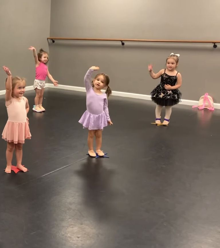 Toddlers in leotards and tutus wave their arms during a dance class in a studio with a black floor and a barre